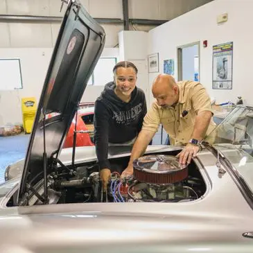 Here's an alt tag for the image: Mechanic and student working on classic car engine.