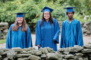 Three graduates in blue gowns and caps standing next to a pile of rocks.