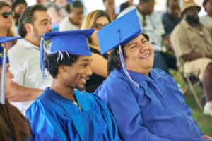 Two graduates are smiling while sitting in a crowd.