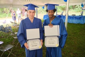 Two young men holding up diplomas in front of a tent.